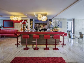 A modern bar area with a red bar counter, multiple stools, and a variety of beverages and fruits displayed. - Novotel Roma Eur (Rome)