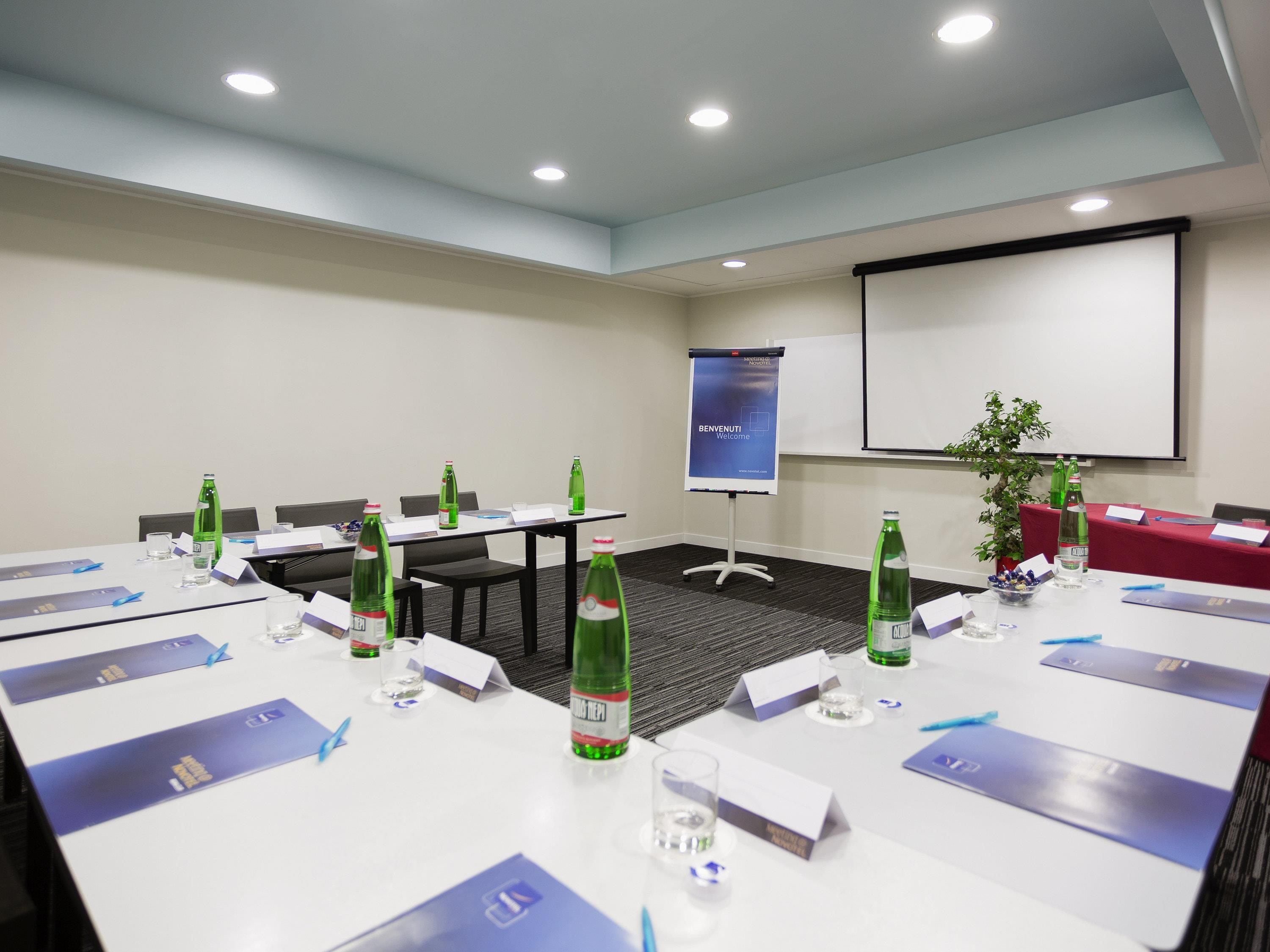 A conference room with tables, chairs, nameplates, water bottles, a projector screen, and a presentation screen.
