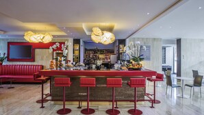 A modern bar area with a red bar counter, multiple stools, and a variety of beverages and fruits displayed. - Novotel Roma Eur (Rome)