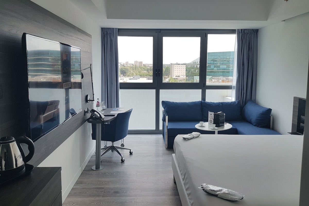 A hotel room with a bed, a desk with a chair, a sofa, and a large window offering a city view.