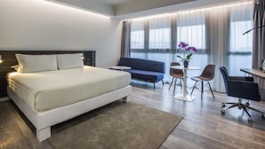 A modern hotel room with a large bed, a sofa, a desk, and a small table with a plant. - Novotel Roma Eur (Rome)