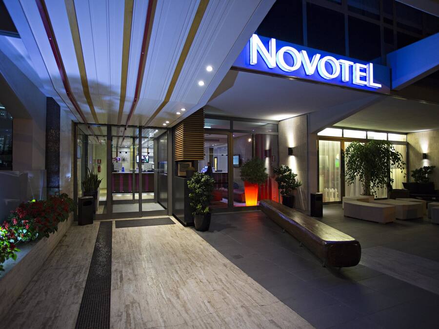 Exterior de un hotel Novotel con diseño moderno, destacando un letrero iluminado, una entrada bien iluminada y zonas de estar al aire libre.