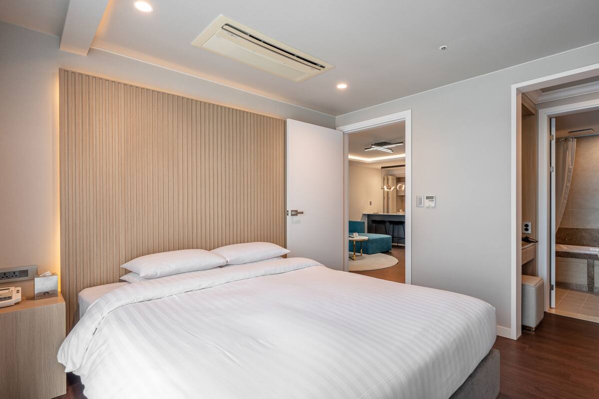 A modern bedroom with a large bed, a bedside table with a lamp, and a wall-mounted air conditioner.