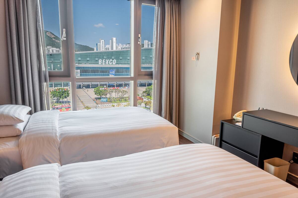A hotel room with two beds, a desk, and a view of a cityscape including a building labeled 'BEXCO'.