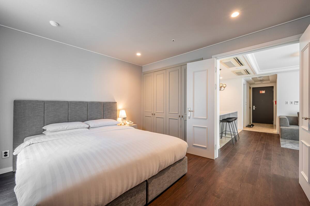 A modern bedroom with a large bed, a nightstand, and a desk with a chair. There are wardrobes and a door leading to another room.