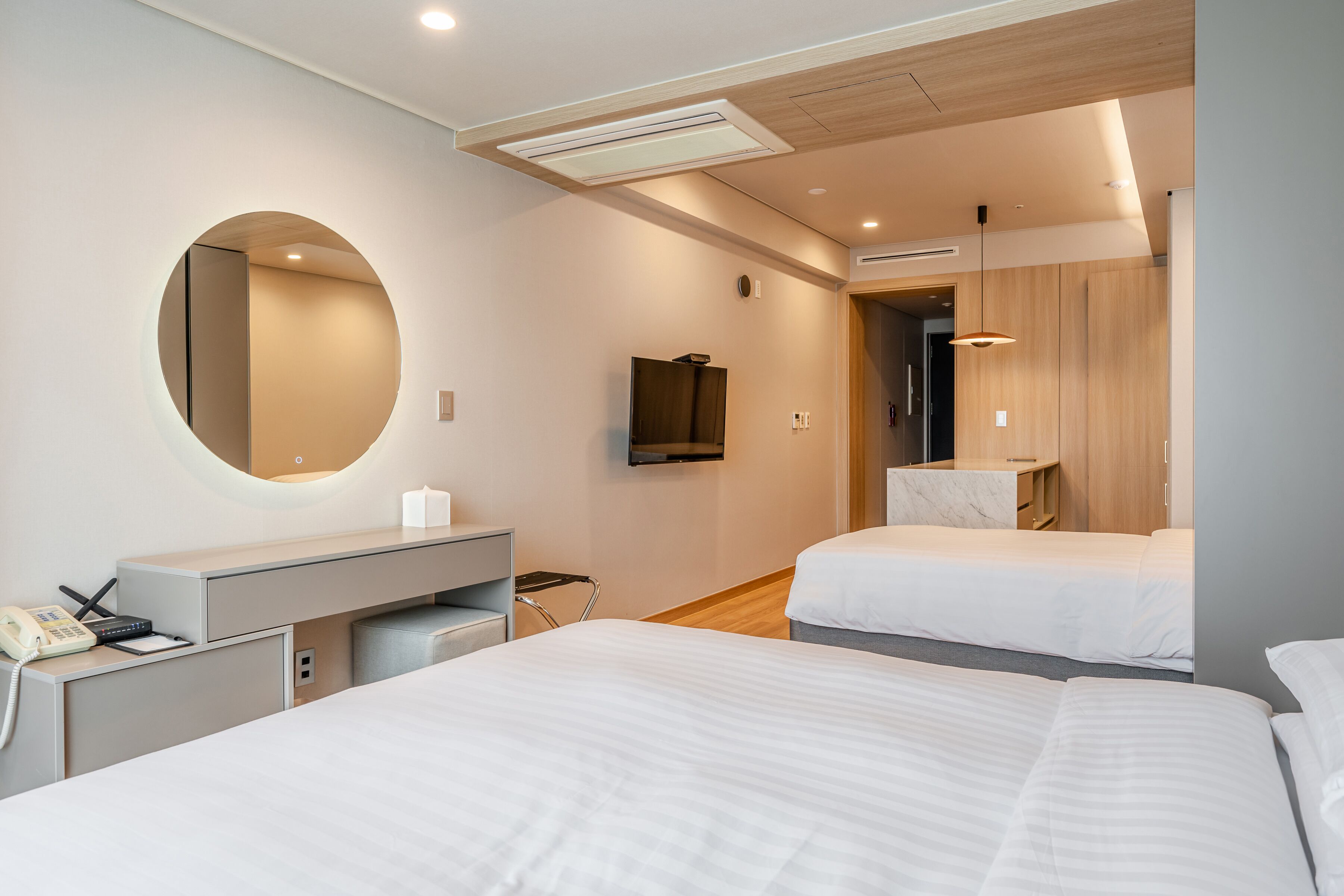 Habitació d'hotel amb dos llits, un escriptori amb telèfon, un mirall rodó, una televisió i un armari de fusta.