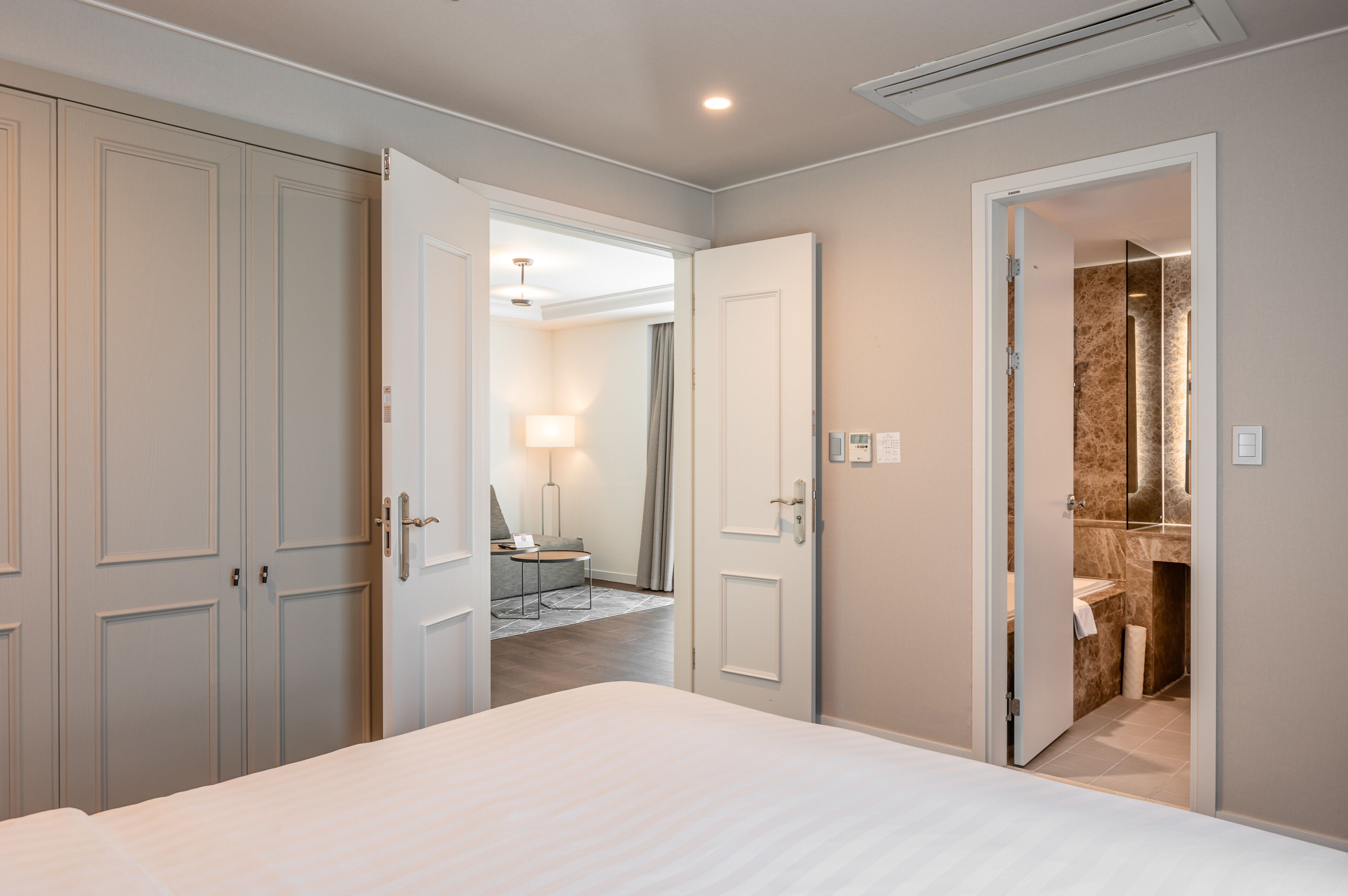 A hotel room with a large bed, a wardrobe, a bathroom with a marble wall, and a small seating area with a lamp.