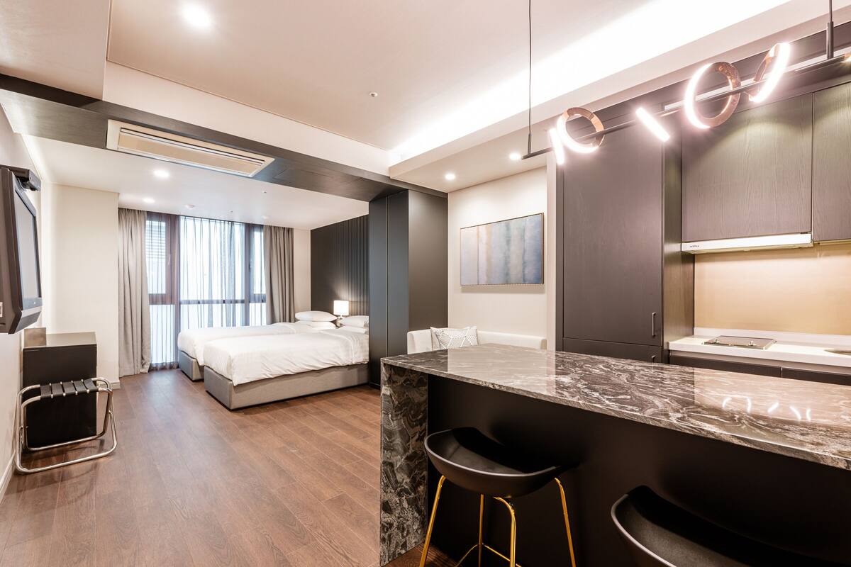A modern hotel room with a marble countertop, black bar stool, a bed with white linens, a large window with curtains, and a minimalist design.