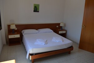 Uma cama bem arrumada com duas almofadas e duas toalhas dobradas, uma cabeceira de madeira, duas mesas de cabeceira com abajures e um pequeno quadro na parede.