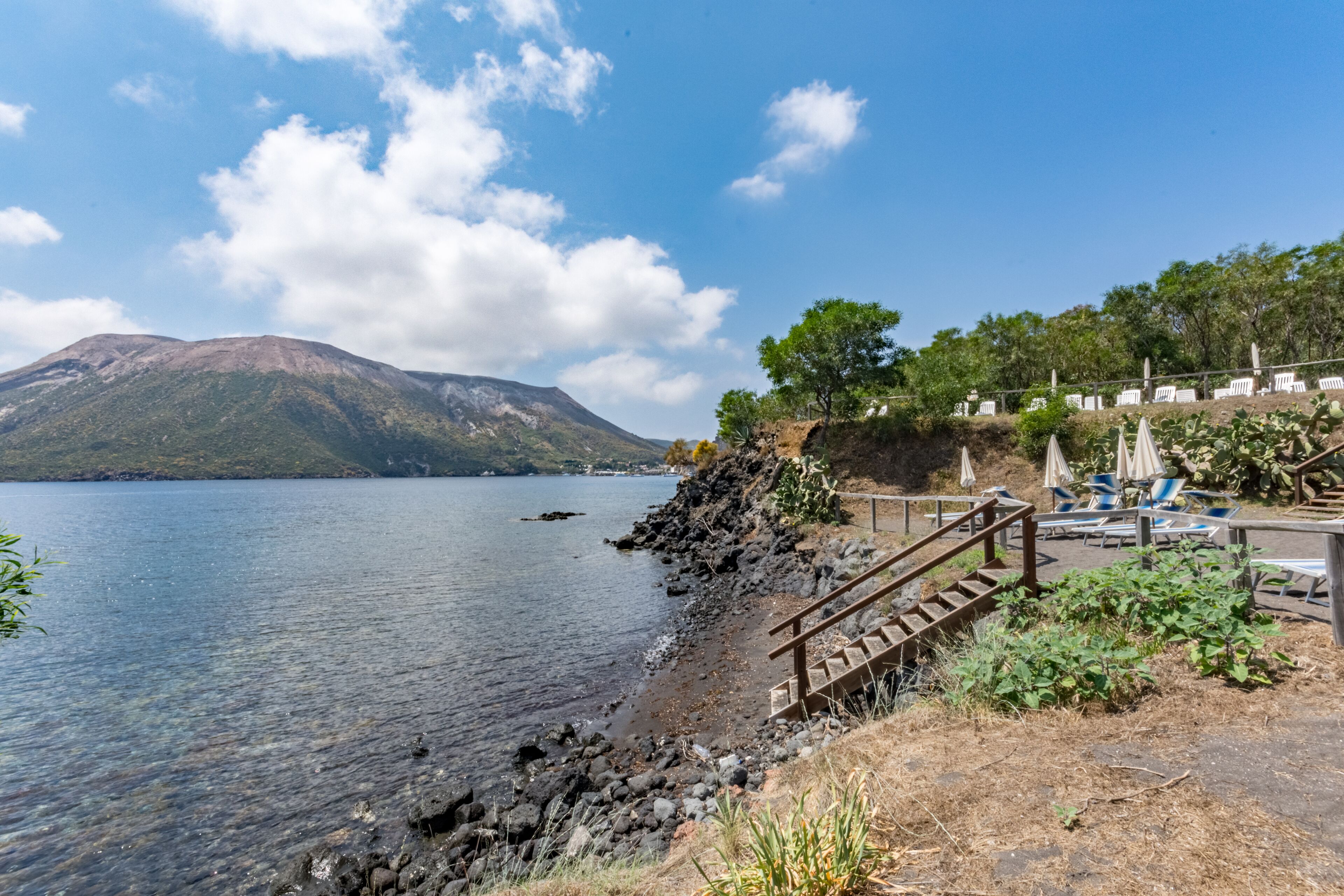 Vista del lago con una montaña al fondo, una escalera de madera que desciende hacia el agua y una hilera de sillas y sombrillas de playa.