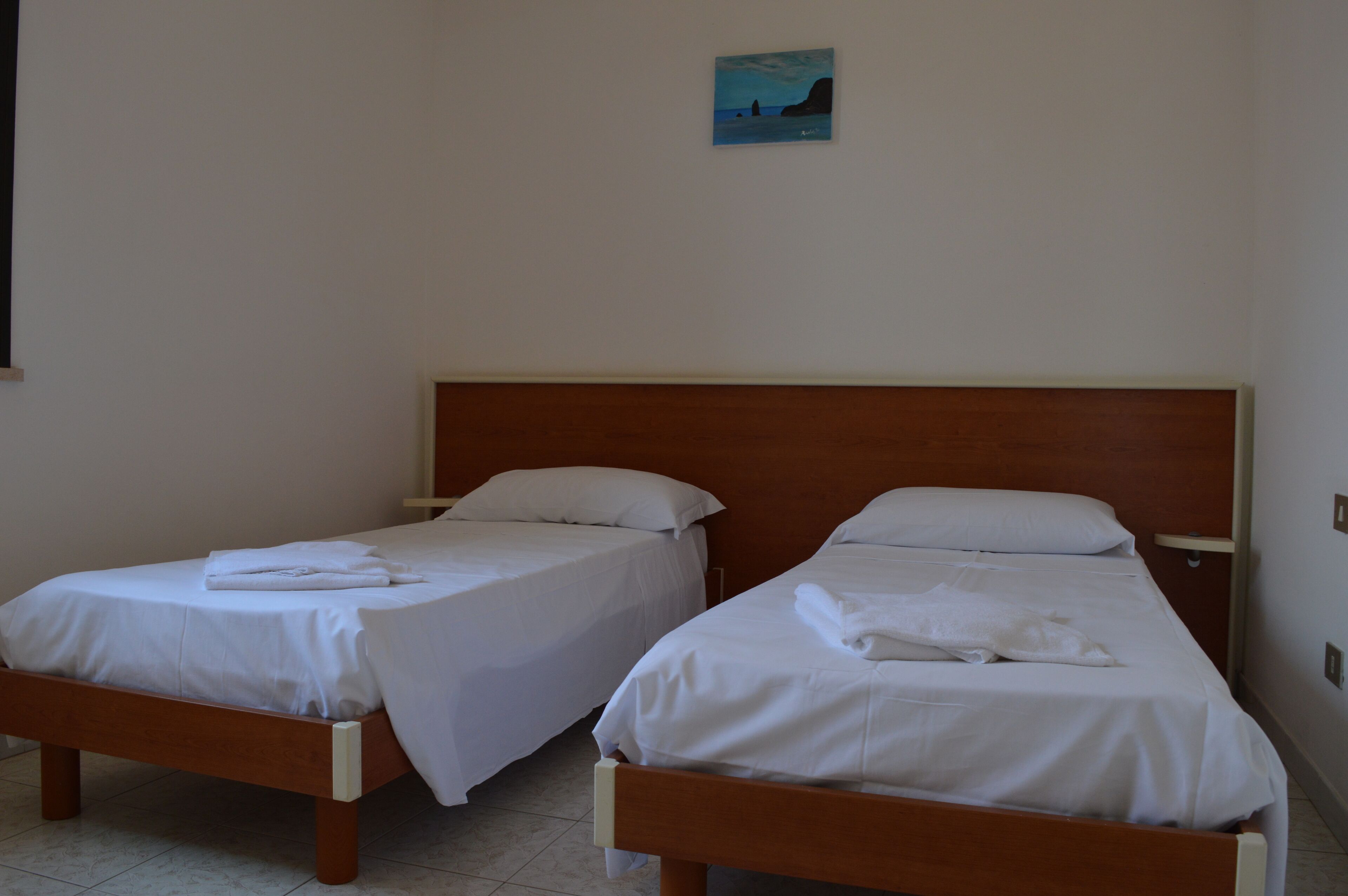 Two single beds with wooden frames and white bedding in a room with white walls and a small painting on the wall.
