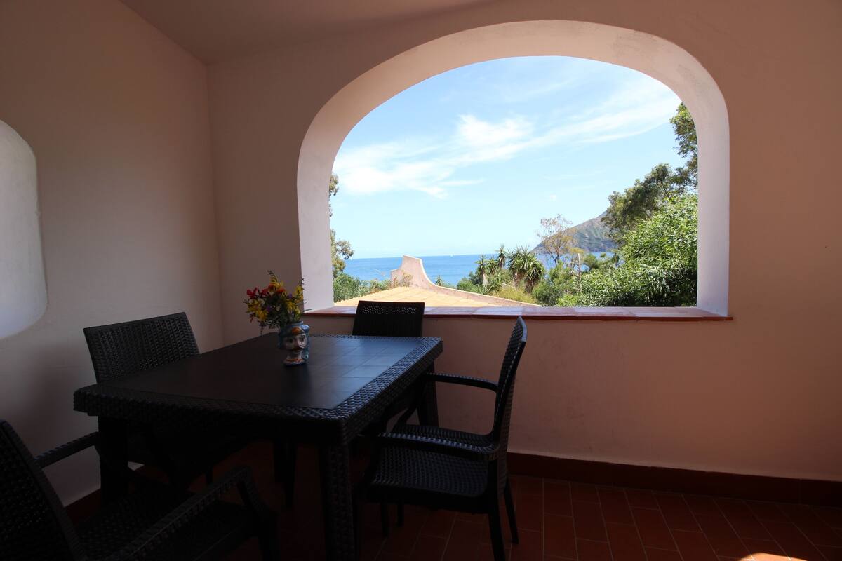Un coin repas avec une table et des chaises, une fenêtre en arc de cercle et une vue sur la mer et les collines.