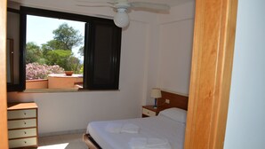 Un dormitorio con cama, mesita de noche, cómoda, ventana con vistas a árboles y flores, y ventilador de techo.
