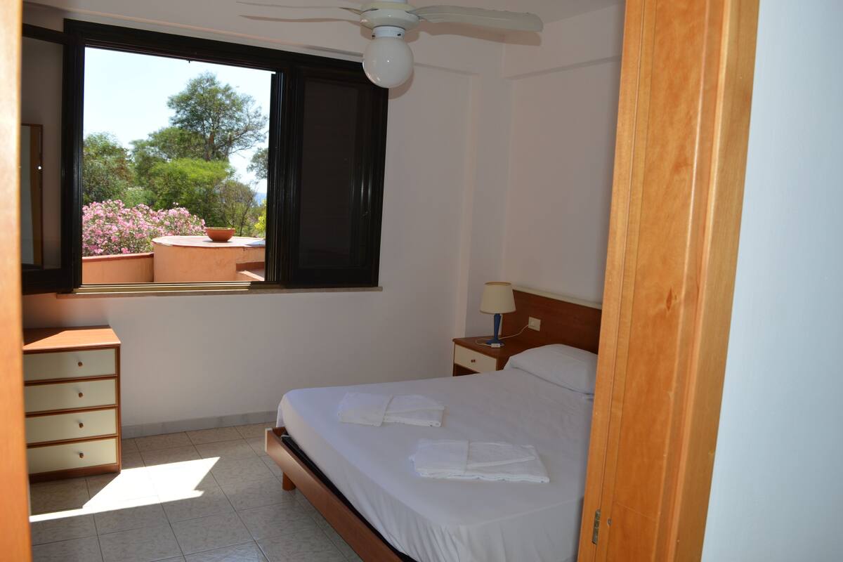 Une chambre à coucher comprenant un lit, une table de chevet, une commode, une fenêtre donnant sur des arbres et des fleurs, et un ventilateur de plafond.