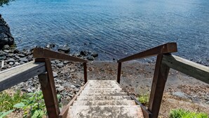 Una escalera de madera que desciende hacia una costa rocosa, con vistas a aguas tranquilas y colinas distantes.