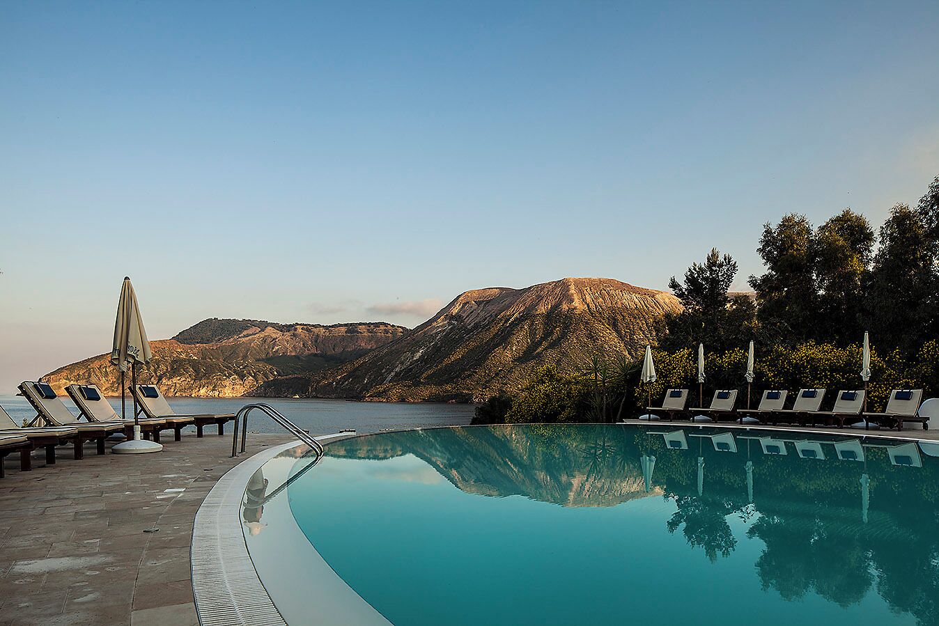 Ein Pool mit Liegestühlen und Sonnenschirmen, umgeben von Bergen und Bäumen.