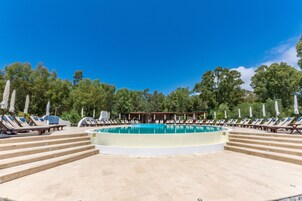 A pool area with lounge chairs and umbrellas, surrounded by trees and a building in the background.