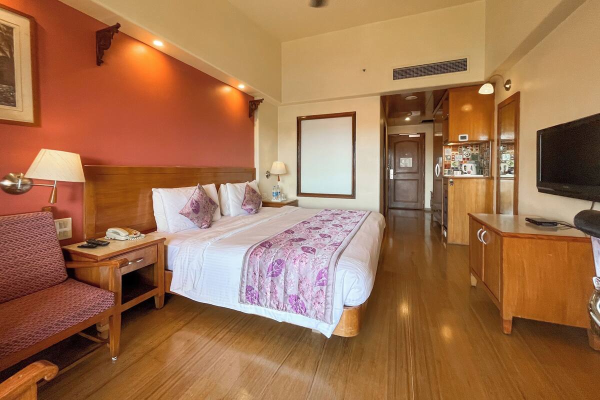 A hotel room with a bed, bedside table, lamp, chair, TV, and a hallway leading to a bathroom.