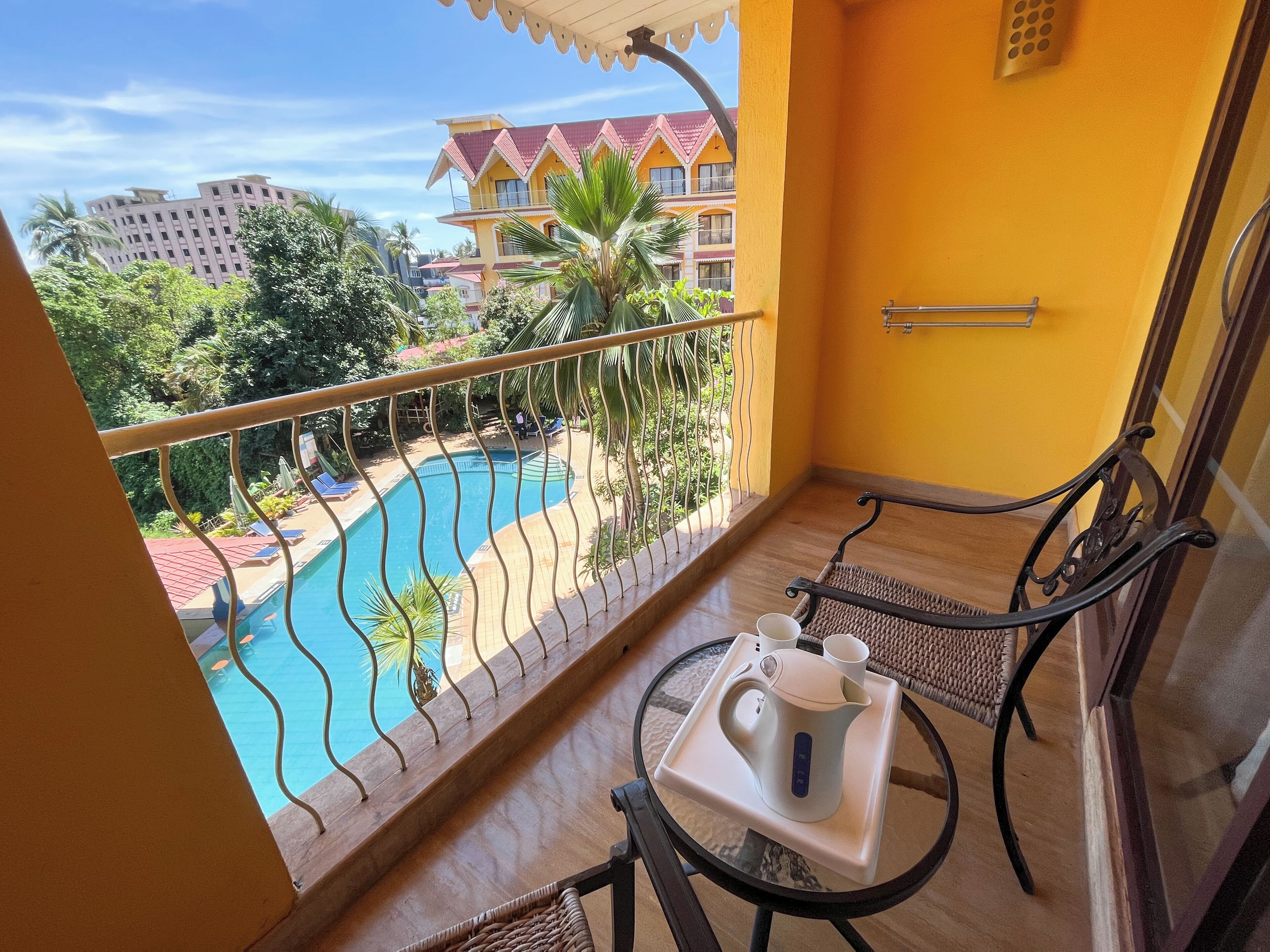 A balcony with a view of a swimming pool, a table with a kettle and cups, and a chair.