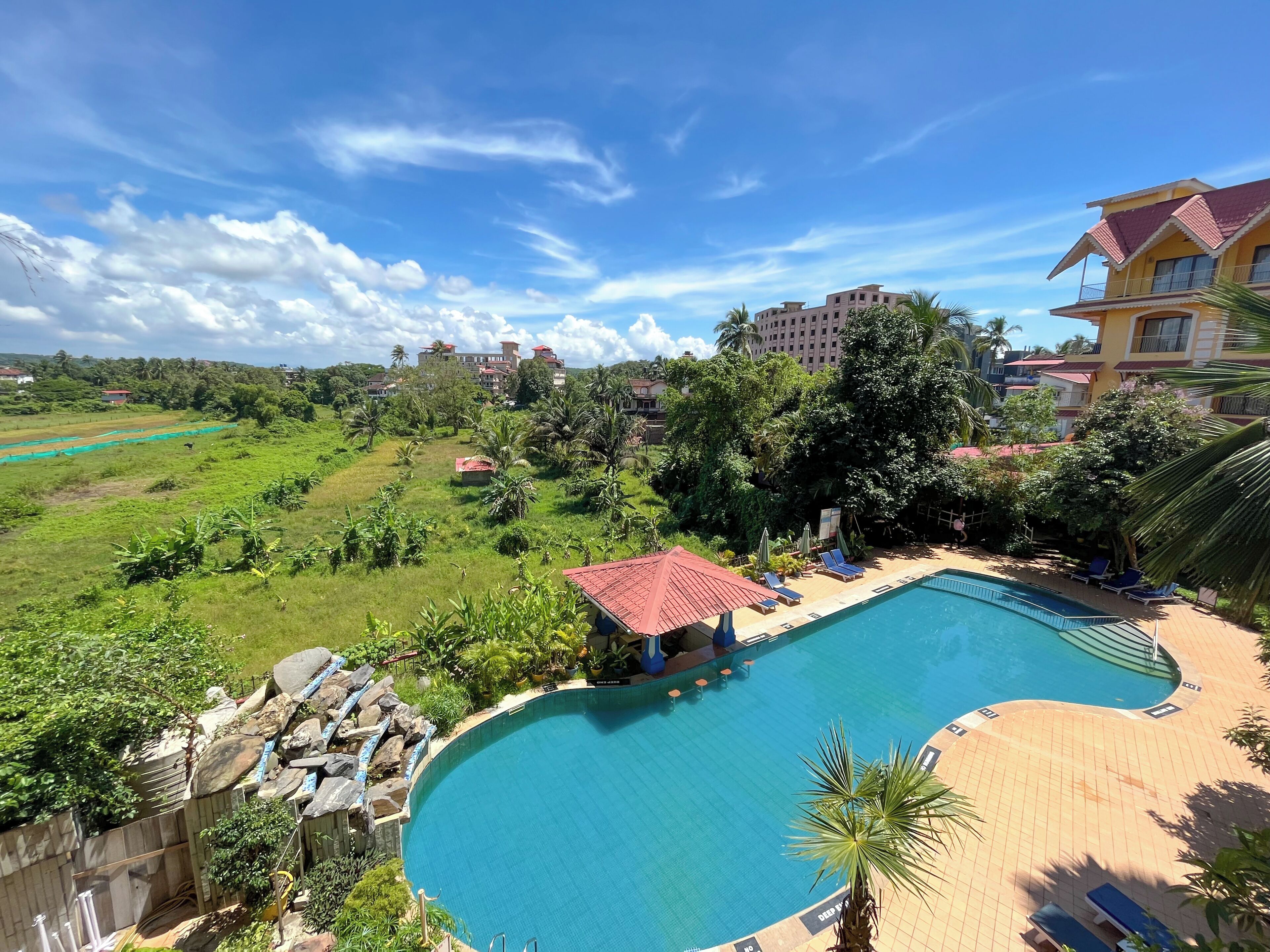 A swimming pool with lounge chairs and a red-roofed structure, surrounded by greenery and buildings.
