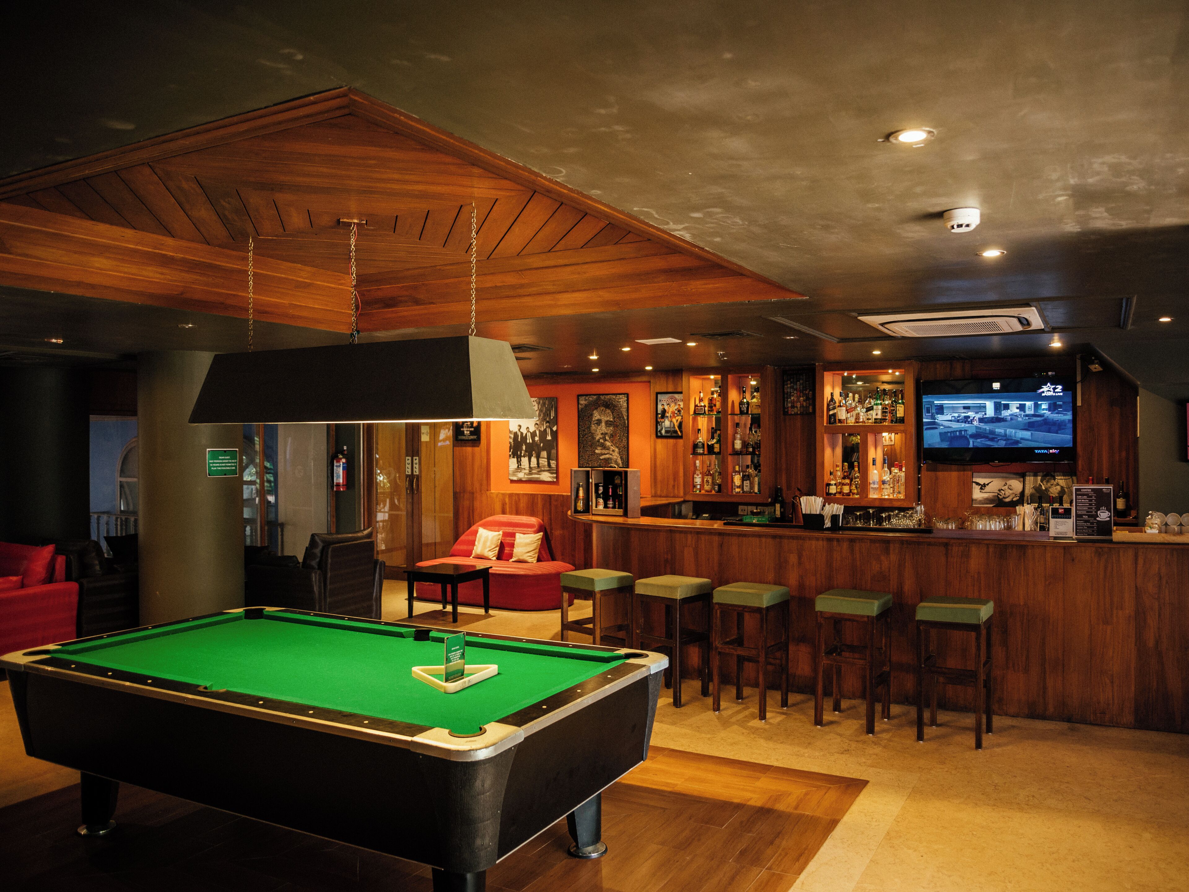 A pool table, bar area, and seating in a room with wooden ceiling and walls.
