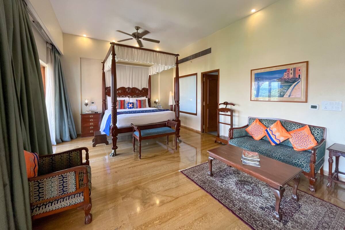 A spacious bedroom with a four-poster bed, a sitting area with a sofa and armchairs, a wooden coffee table, and a large window with curtains.