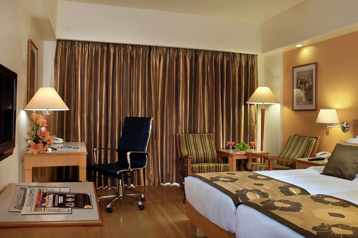 A hotel room with a bed, two chairs, a desk with a lamp, a small table with a drink, and a picture on the wall.