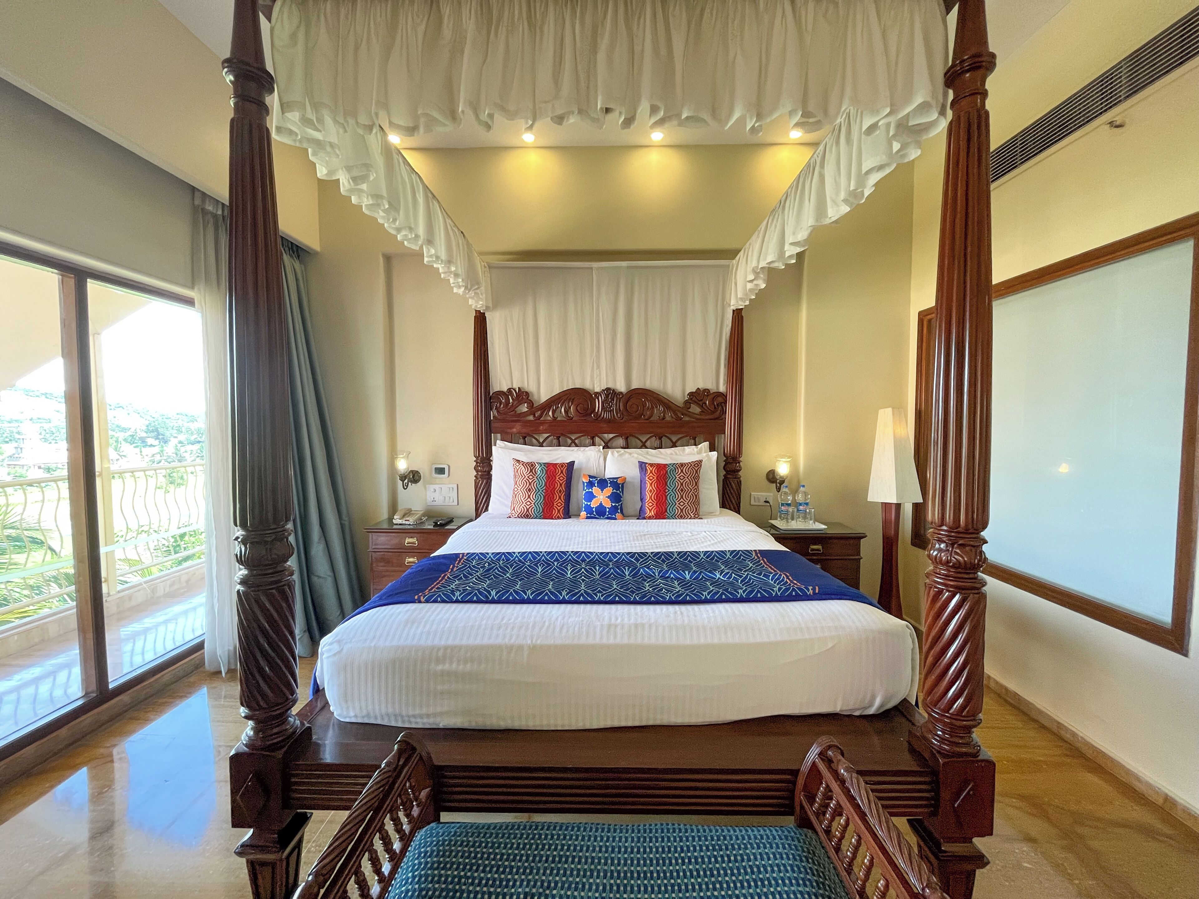 Una cama con dosel y cuatro columnas, con cubrecama blanco y almohadas de colores, una mesita de noche con lámpara y vistas al verdor exterior.