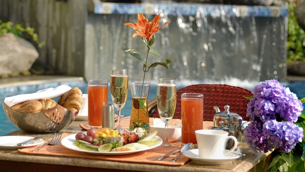 A breakfast setup with croissants, fruit, and beverages by a pool with a waterfall backdrop. - Acron Candolim Regina (Candolim)