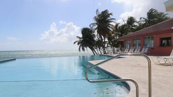 A clear blue pool with a metal ladder, palm trees, and a building with a red roof.