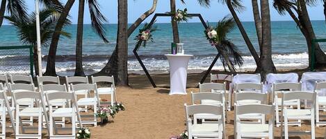 Montaje para una boda en la playa, con sillas y una mesa blancos, decorados con flores.