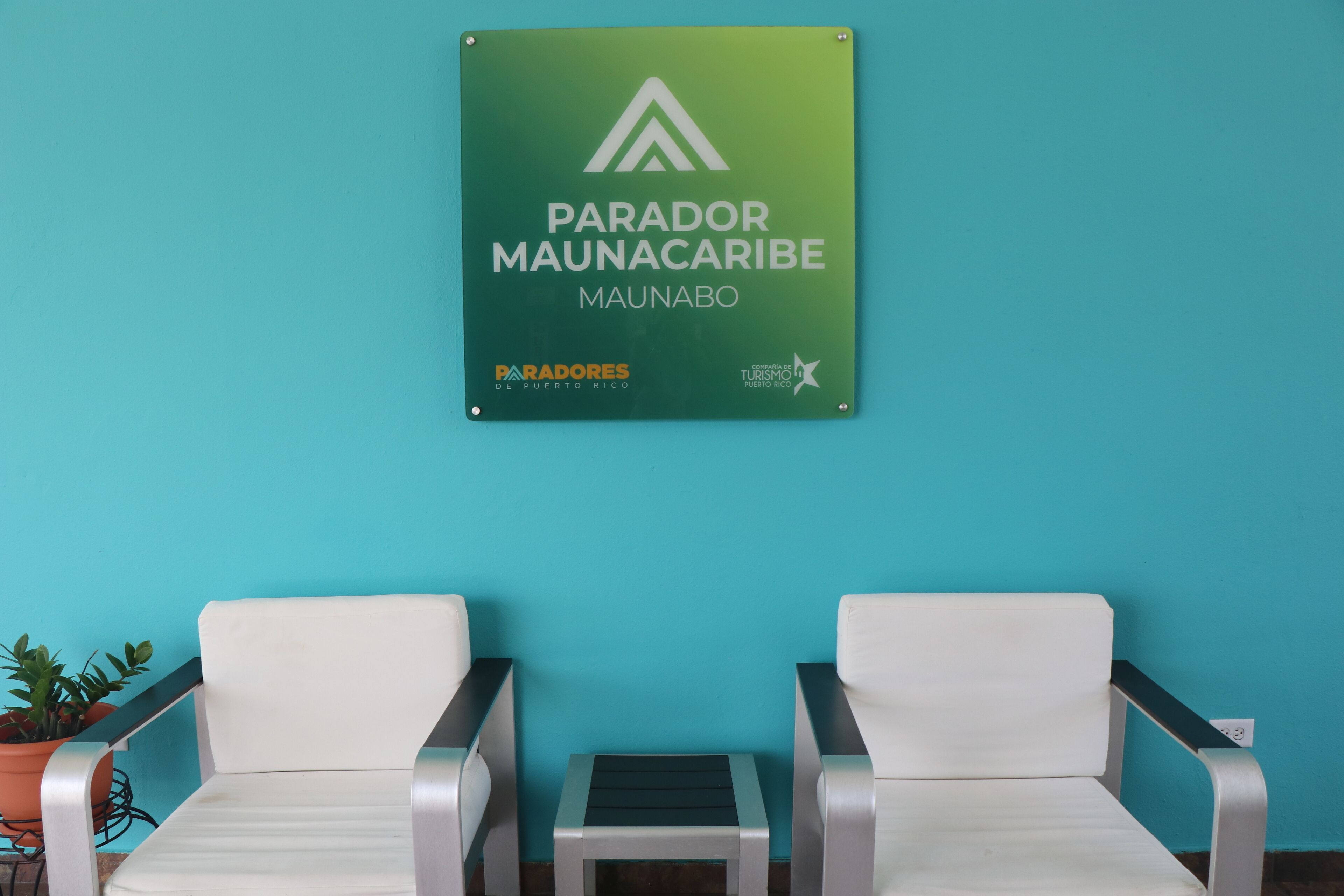 A sign for 'Parador Maunaco' with two white chairs and a small table against a turquoise wall.