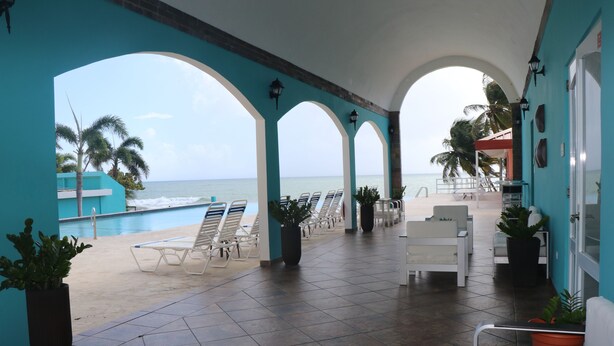 A covered outdoor area with a view of the ocean, featuring lounge chairs, potted plants, and a tiled floor. - Parador MaunaCaribe (Maunabo)
