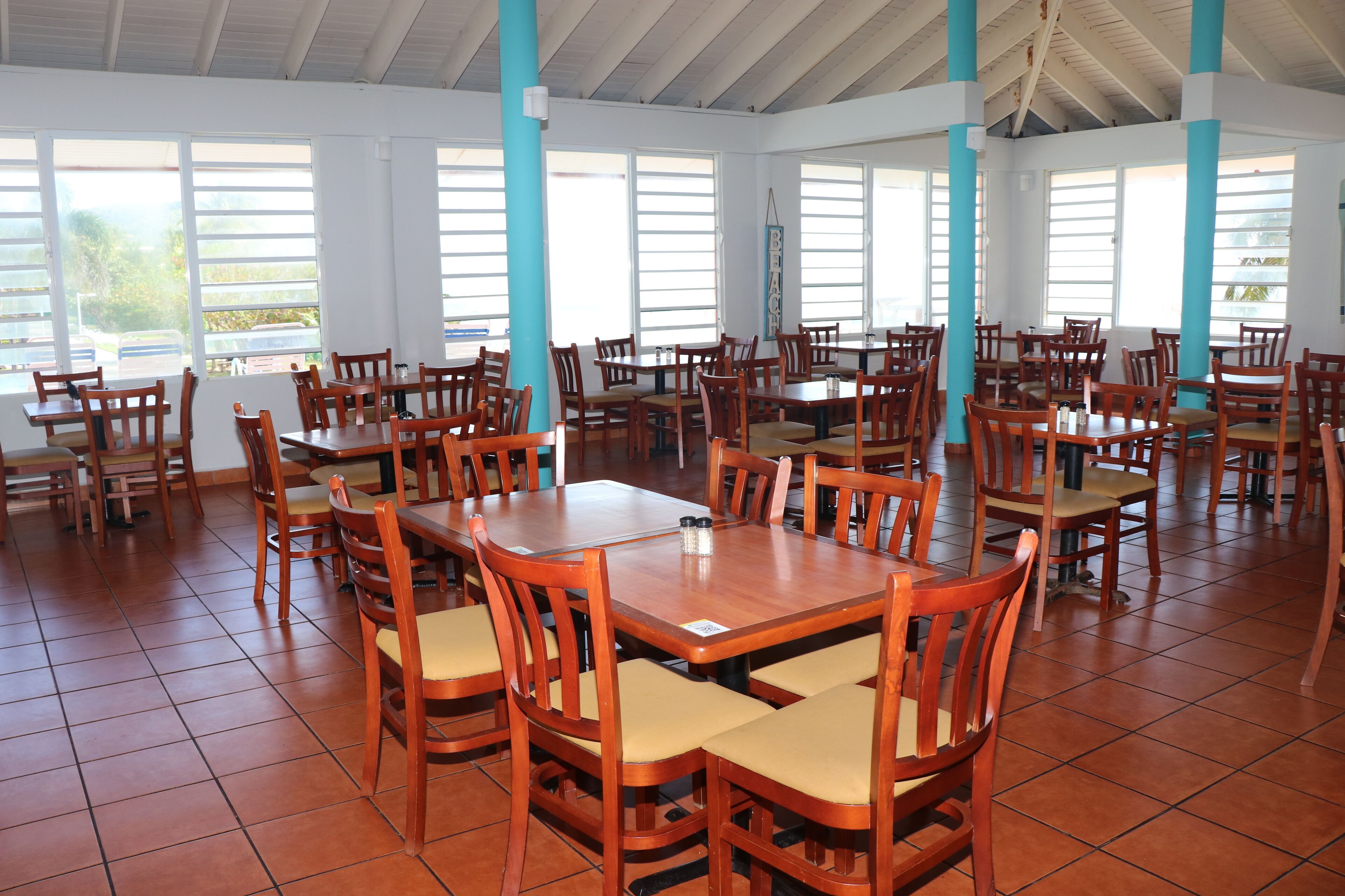A spacious dining area with multiple wooden tables and chairs, large windows, and a tiled floor.