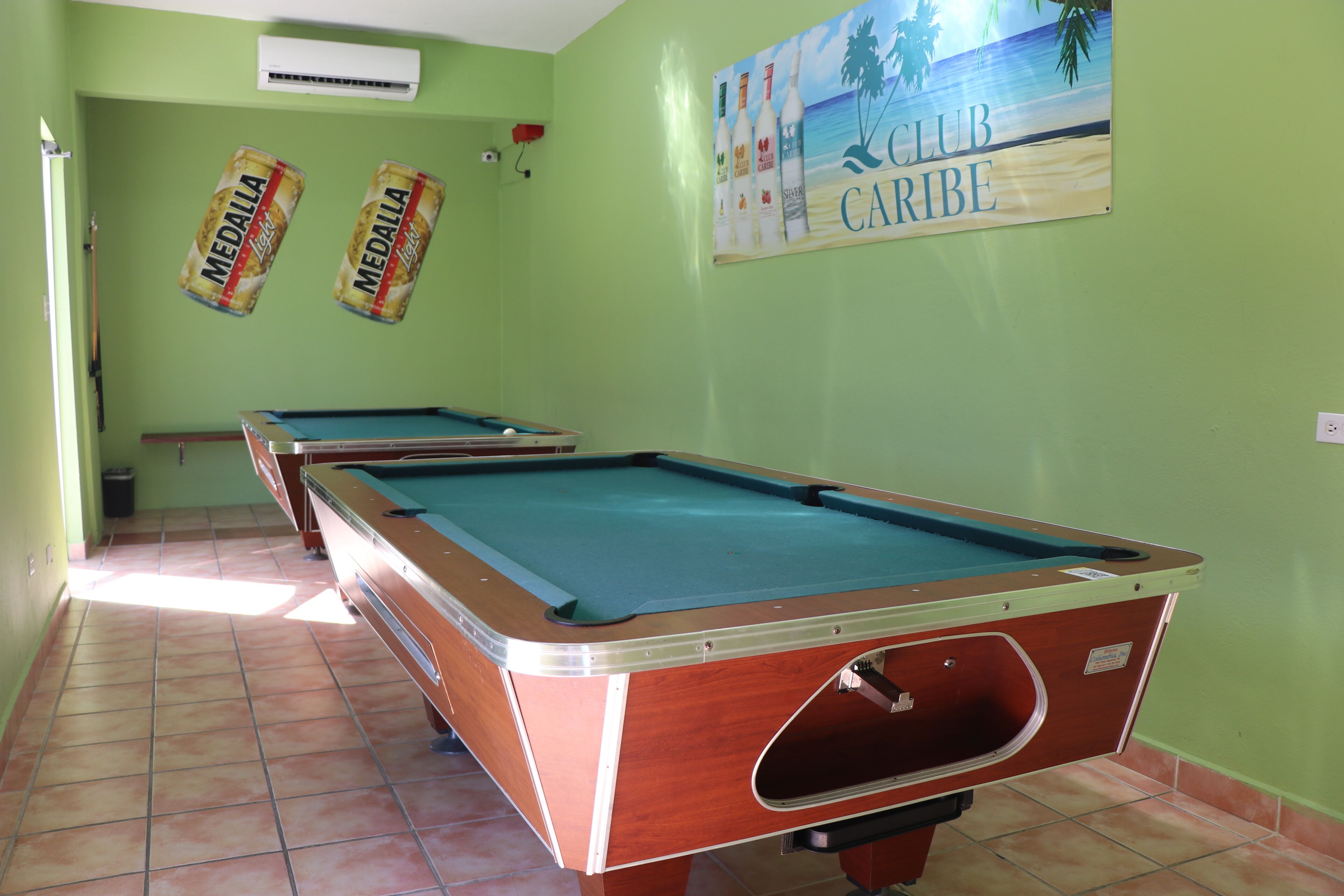 A pool table in a room with green walls, two Medalla beer cans on the wall, and a 'Club Caribe' sign.