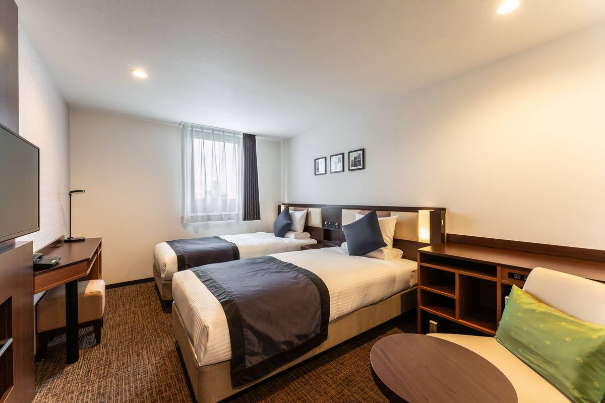 Ein Hotelzimmer mit zwei Betten, einem Schreibtisch, einem Stuhl, einem Fernseher und einem fenster mit Vorhängen.
