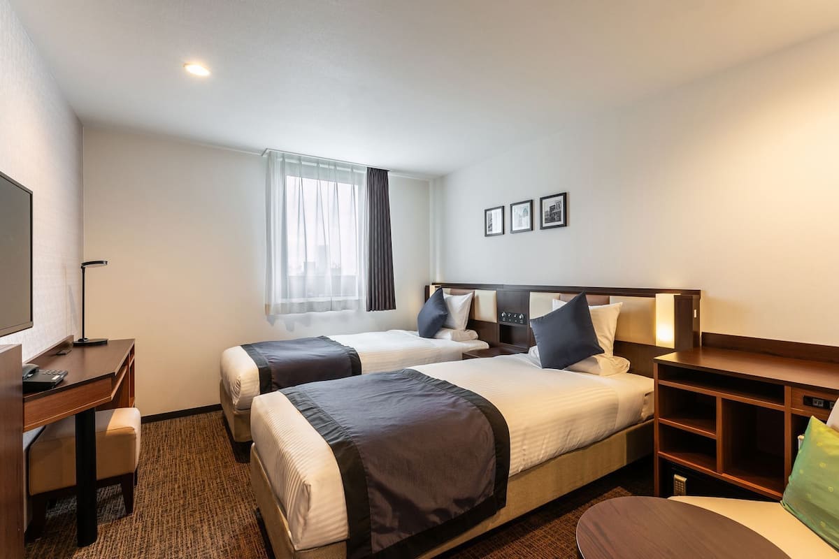 Ein Hotelzimmer mit zwei Betten, einem Schreibtisch, einem Stuhl, einem Fernseher und einem Fenster mit Vorhängen.