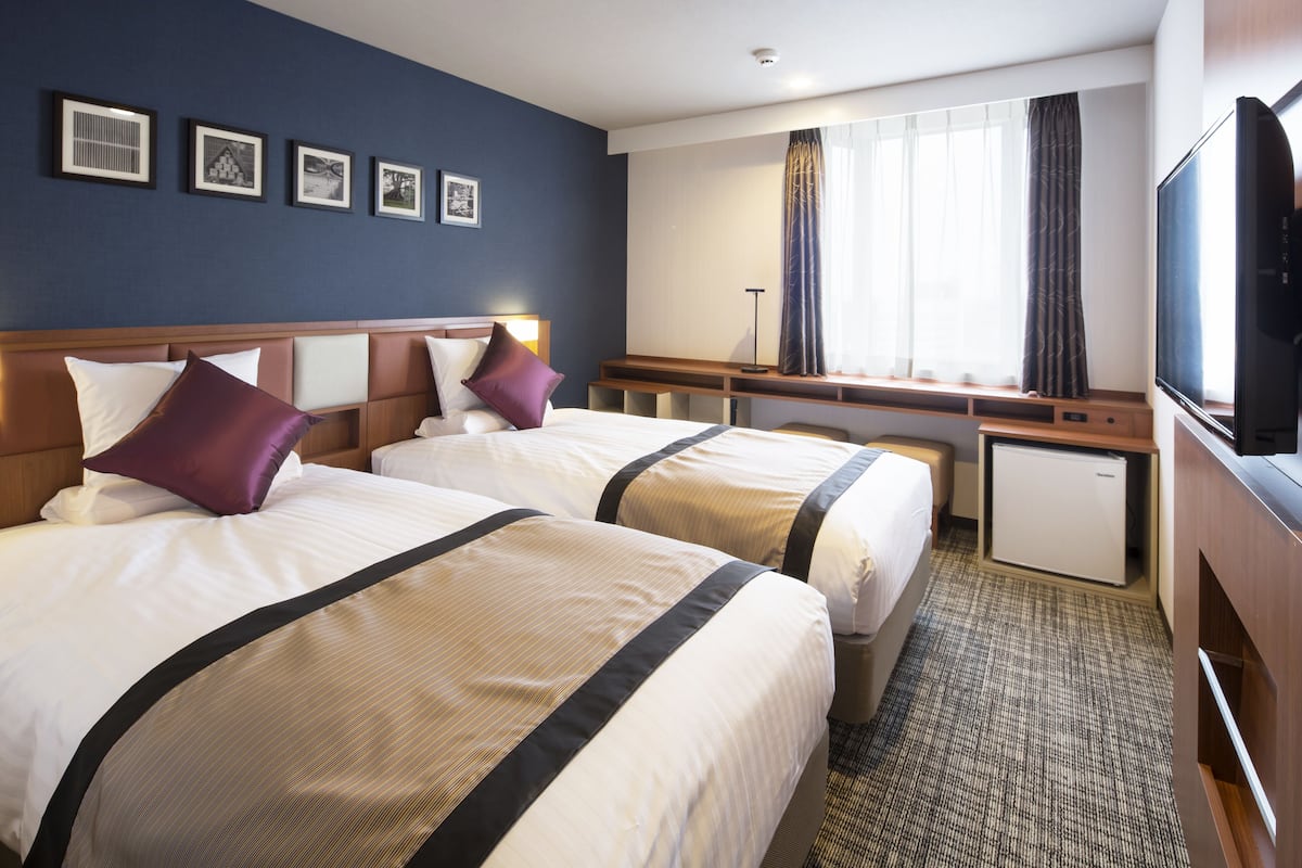 Ein Hotelzimmer mit zwei Betten, einem Schreibtisch, einem Fernseher und einem kleinen Kühlschrank.