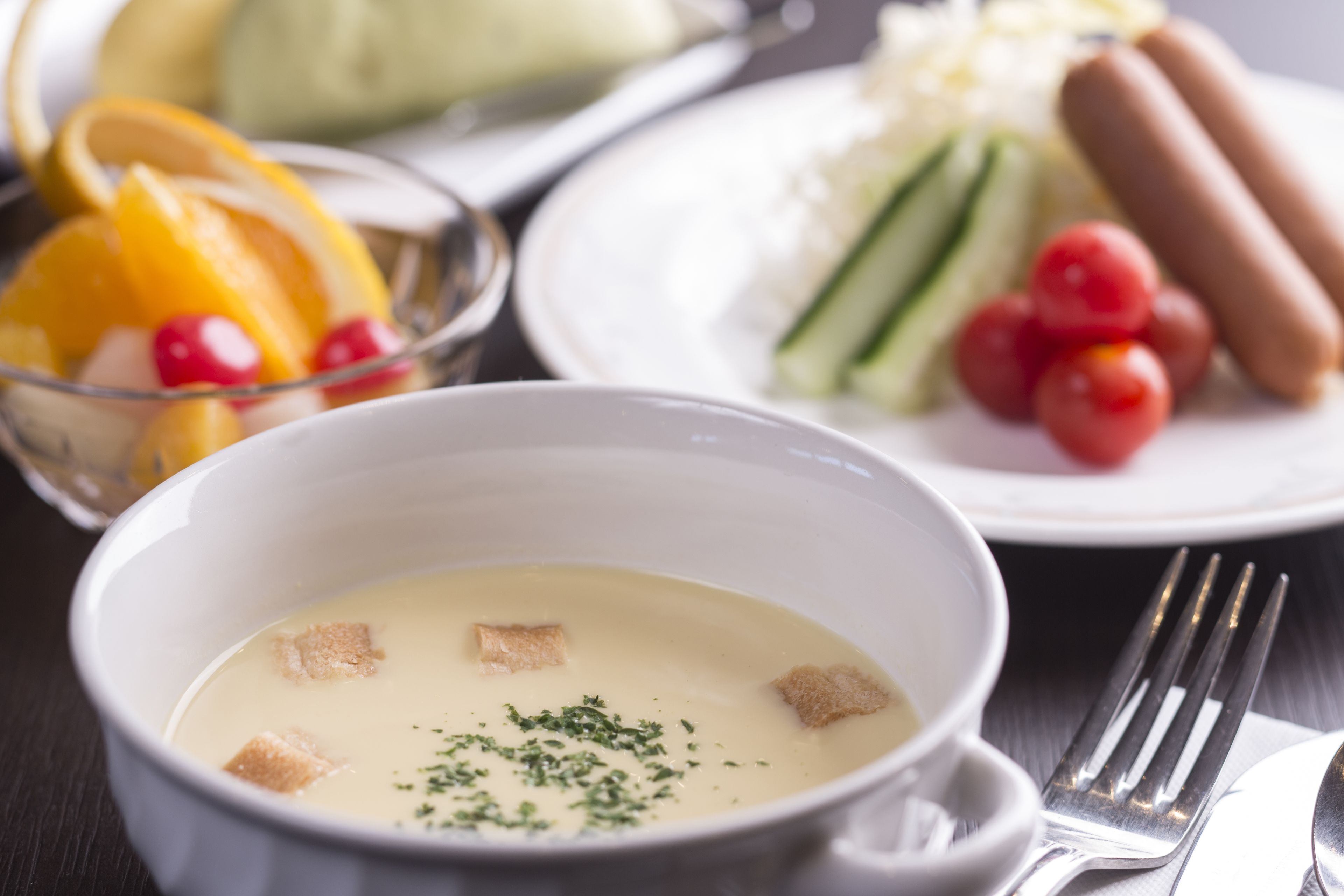 Een kom soep met croutons en kruiden, een bord met worst, groenten en een schaal fruit.