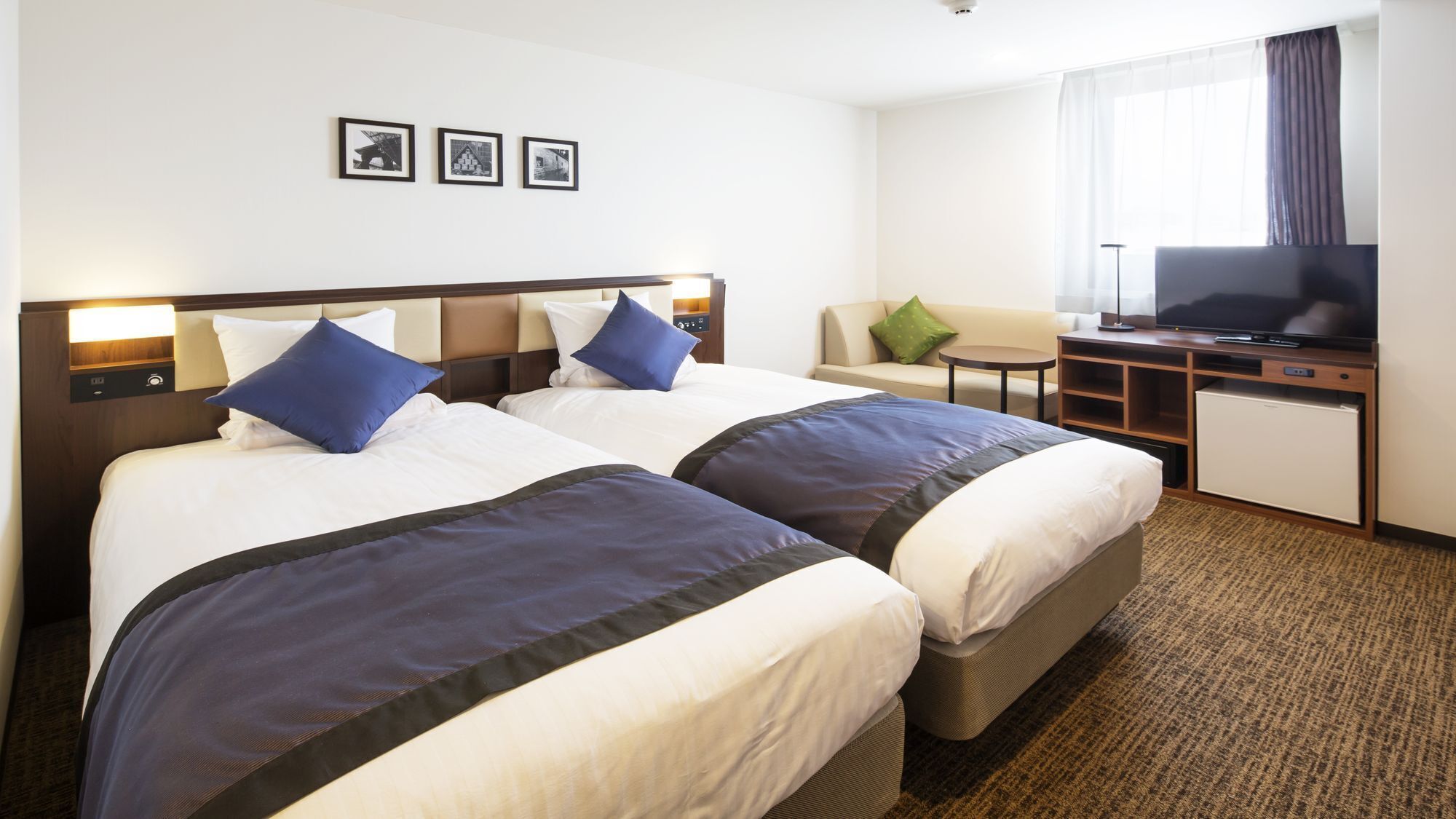 A hotel room with two beds, a desk with a television, a small table, and a window with curtains.
