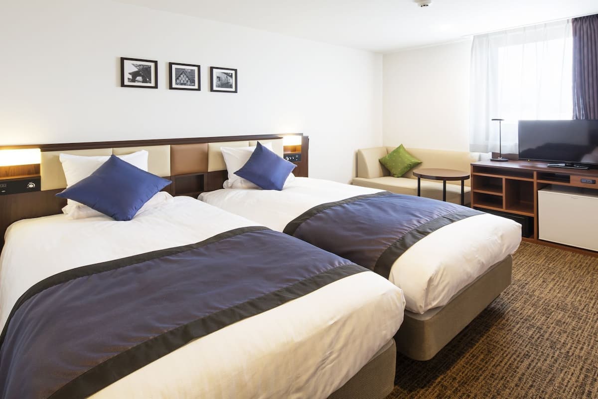 Ein Hotelzimmer mit zwei Betten, einem Schreibtisch mit Fernseher, einem kleinen Tisch und einem Fenster mit Vorhängen.