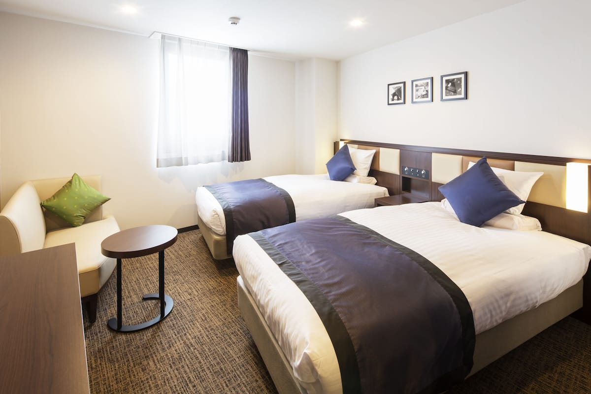 Ein Hotelzimmer mit zwei Betten, einem Schlafsofa, einem kleinen Tisch und einem Fenster mit Vorhängen.