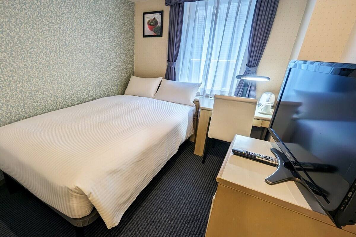 A hotel room with a single bed, a desk with a TV and remote, a chair, and a window with curtains.