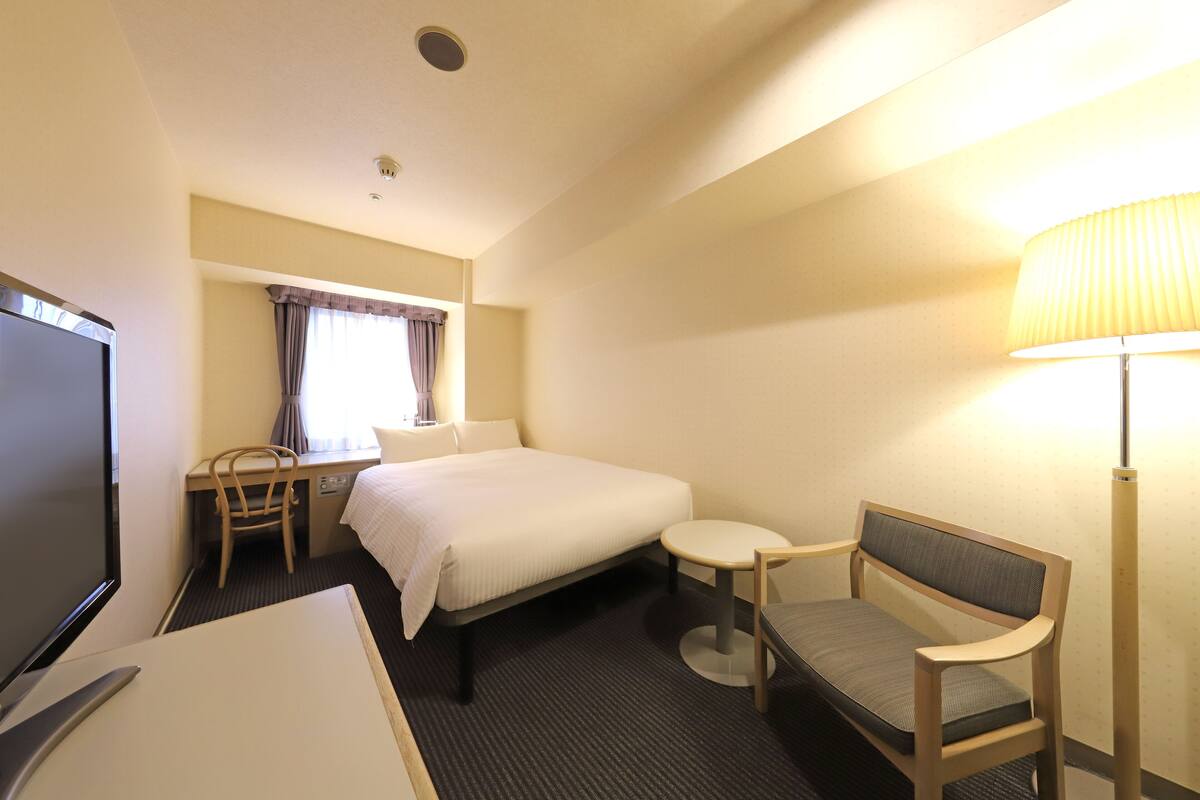A hotel room with a bed, a desk with a chair, a TV, a lamp, and a window with curtains.