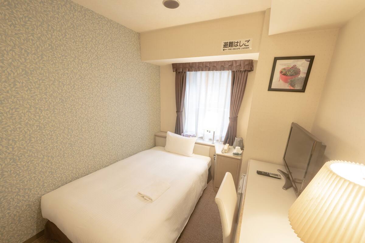 A hotel room with a bed, a desk with a chair, a television, a lamp, and a window with curtains.
