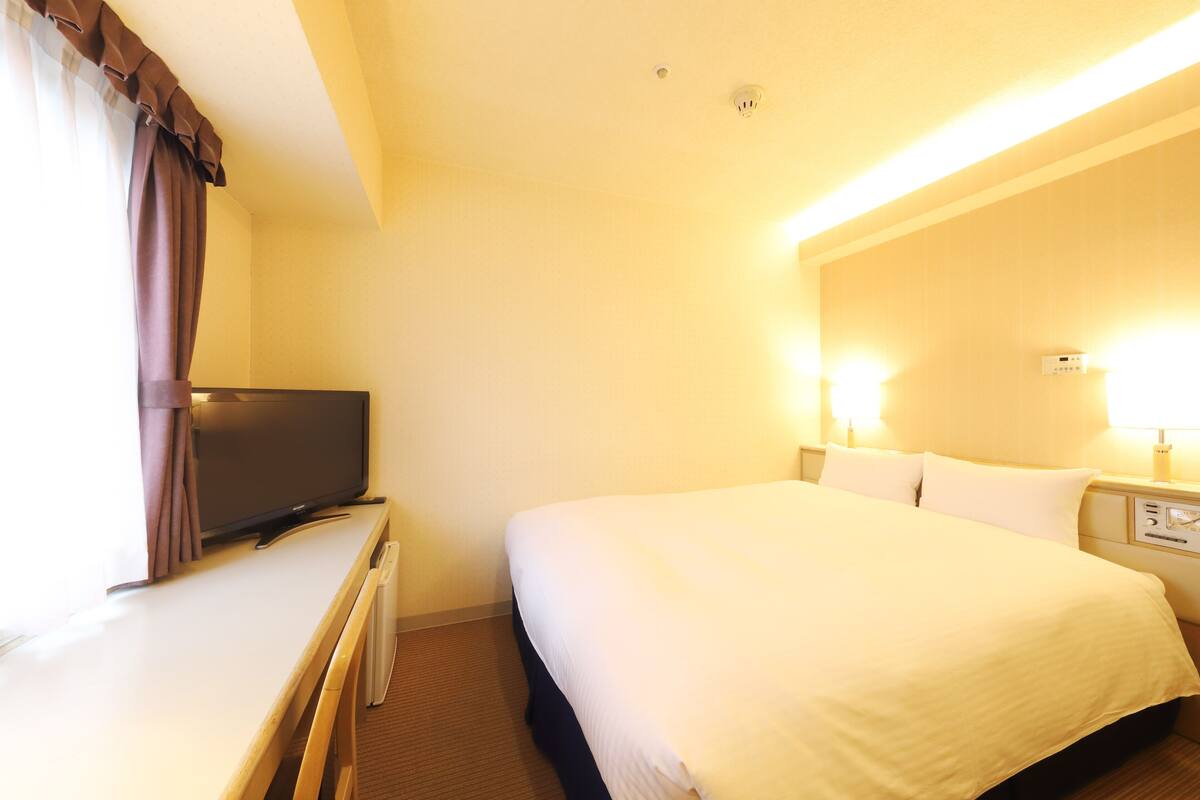 A hotel room with a bed, a television on a desk, and a nightstand with a lamp.
