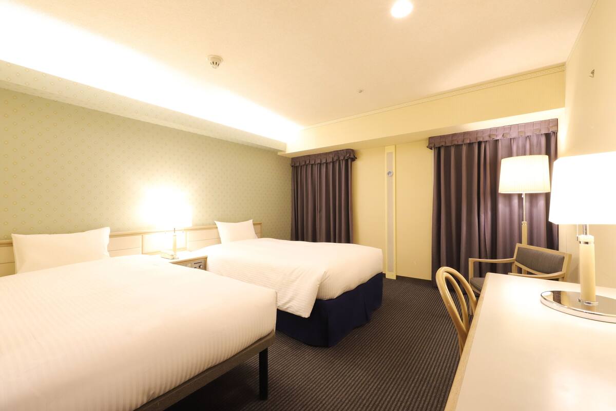A hotel room with two beds, a desk, a chair, and a lamp.