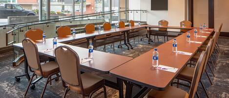 Un salón de conferencias con mesas largas y sillas, equipado con botellas de agua y documentos, ventanas amplias con vistas a la ciudad y una barandilla de cristal.