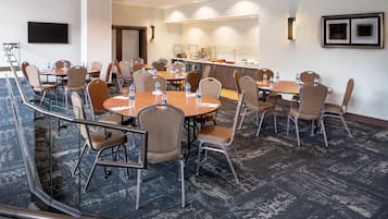 Un salón de conferencias con varias mesas y sillas, equipado con botellas de agua, una televisión de pantalla plana y una zona de bufé.