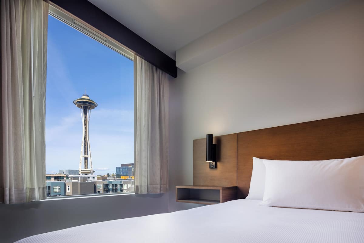 A hotel room with a large bed, a wooden headboard, a nightstand, and a window with a view of a tower and cityscape.